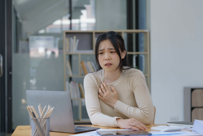 An Asian female has a heart attack pain, tension Sick employee feeling heartache while working on laptop at workplace at desk in office Sad woman in a suit keeps her hand on her chest indoor